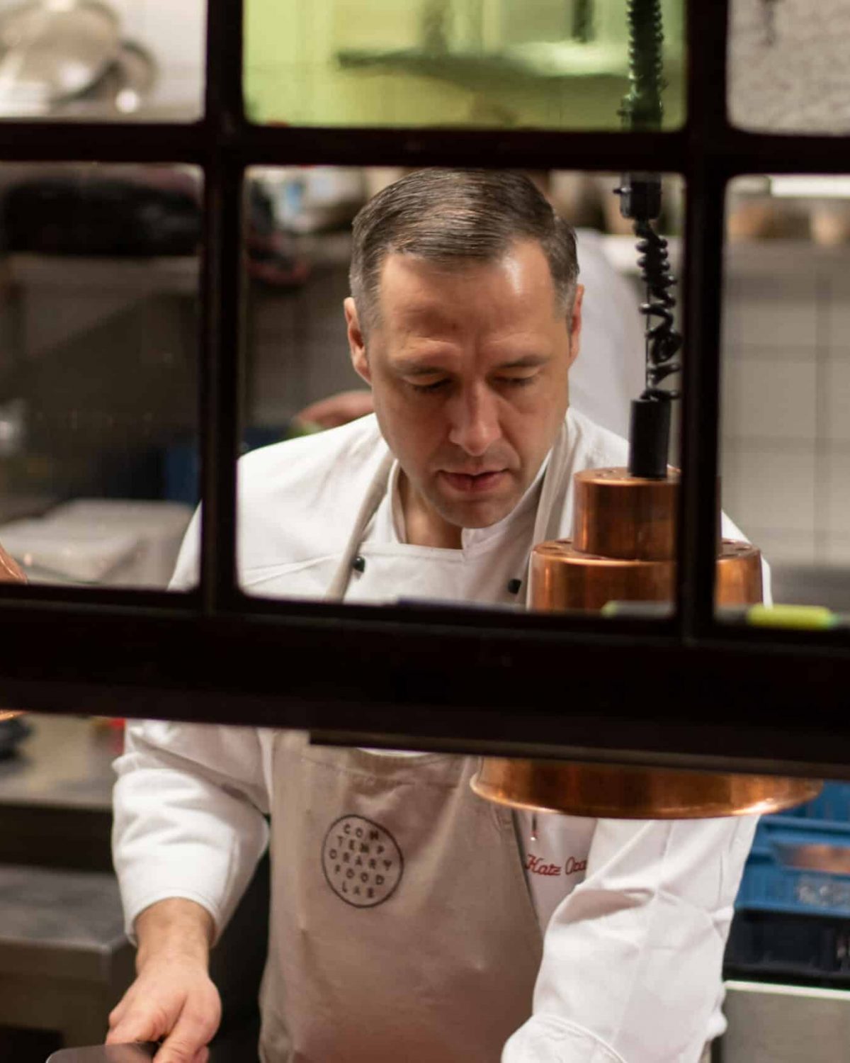 Ein engagierter Koch, fokussiert auf seine Arbeit in einer professionell ausgestatteten Küche, betrachtet durch ein Fenster mit Kupferlampen.