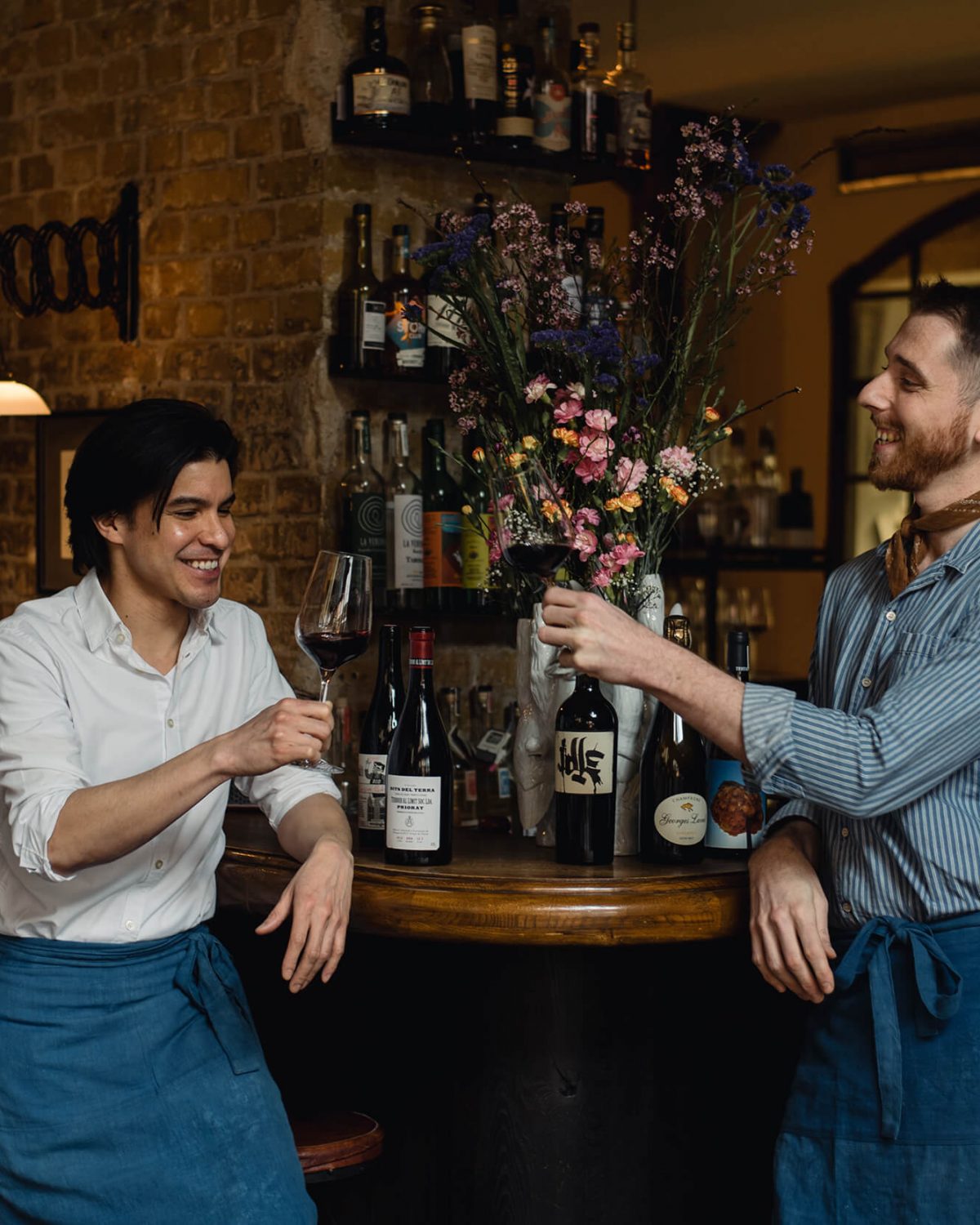 Zwei Männer lachen und stoßen in einer gemütlichen Bar mit Gläsern Rotwein an, umgeben von einer Vielzahl von Weinflaschen und einer mit Blumen befüllten Blumenvase.