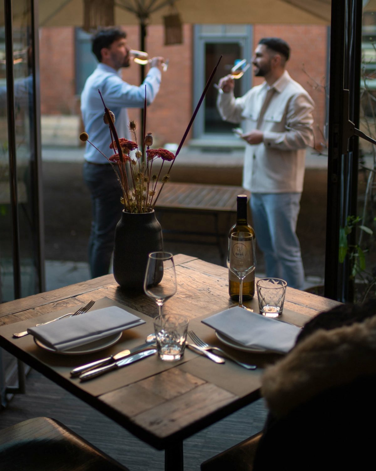 Two men drinking wine outside in dim light with a foreground of a table with a vase and wine bottle.